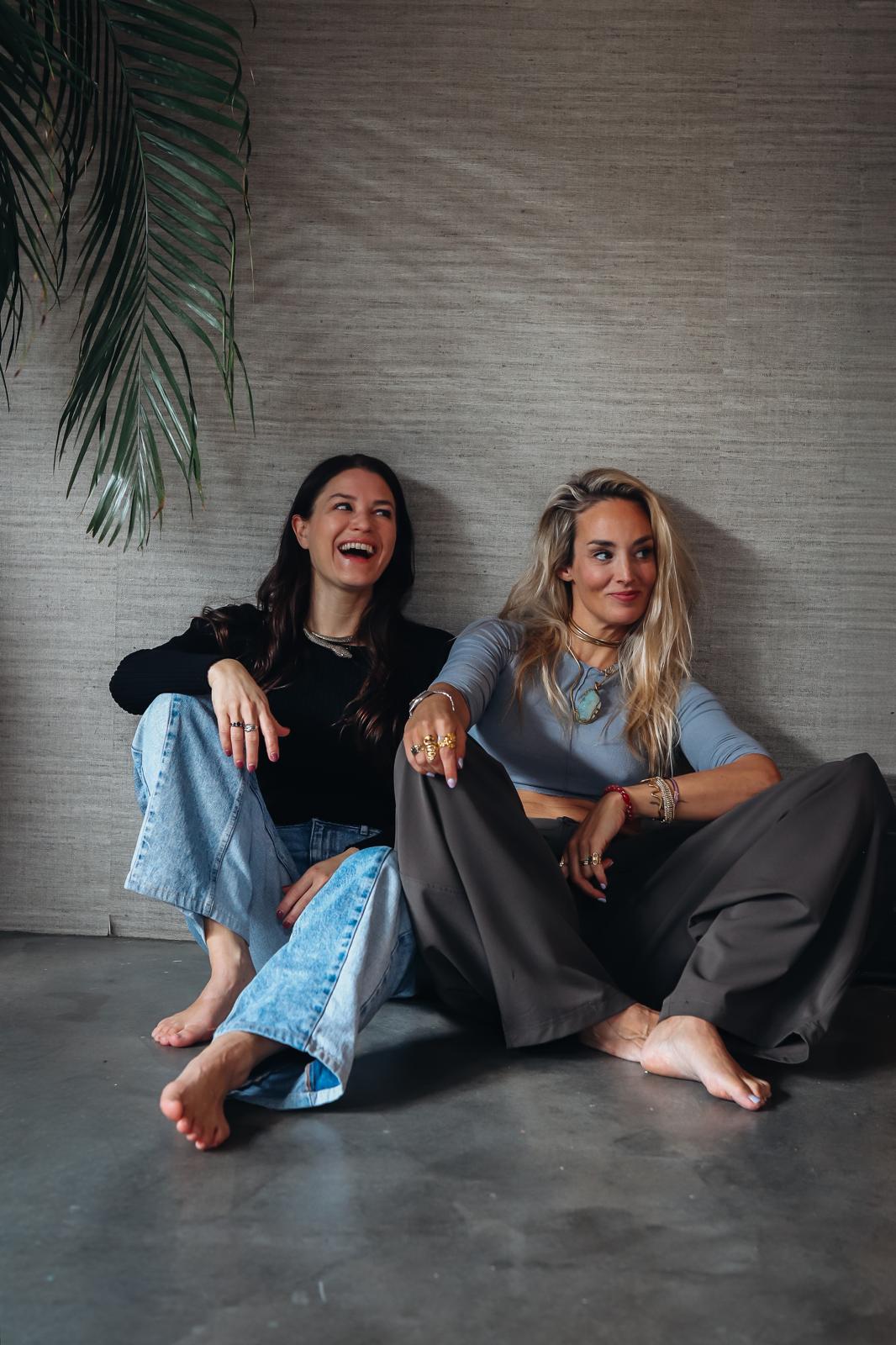 Amanda en Eline zitten lachend tegen een muur, met palmbladeren linksboven
