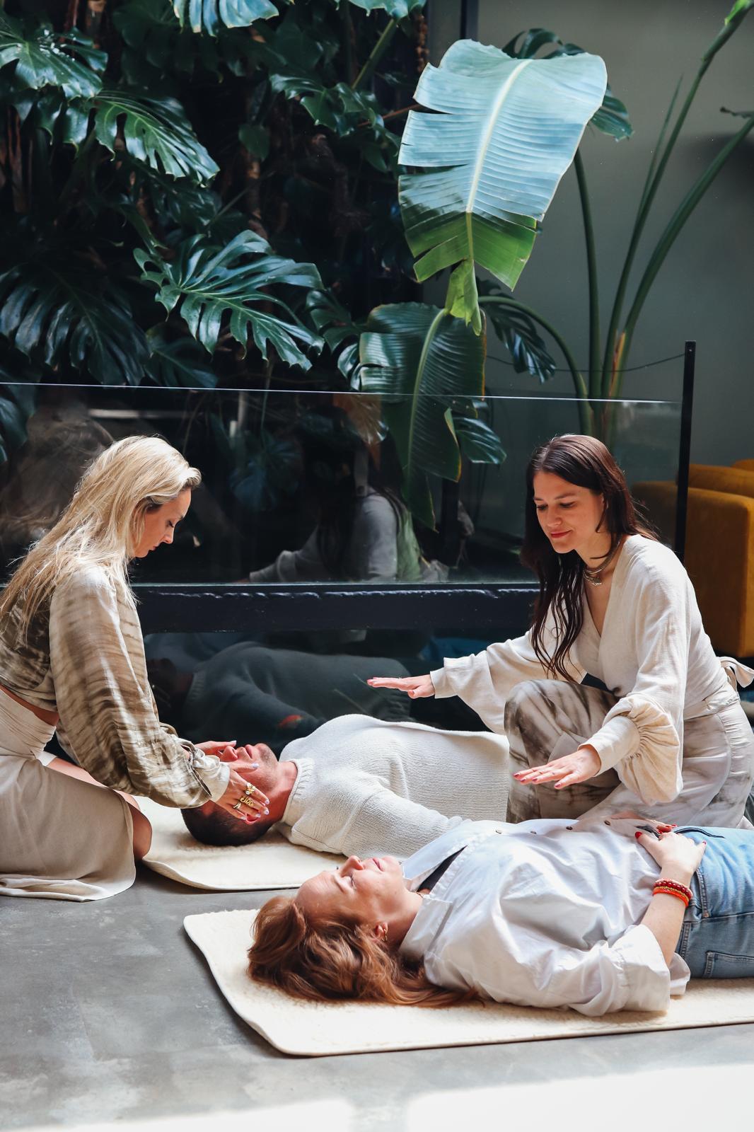 Amanda en Eline begeleiden twee deelnemers bij een sessie, met een groot palmblad op de achtergrond